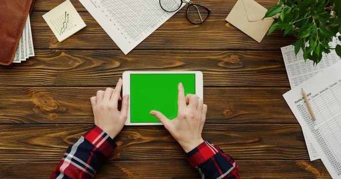 Close up of the female hands with the red motley sleeves working on the white tablet device with a green screen horizontally on the wooden office table with office stuff and money in the wallet