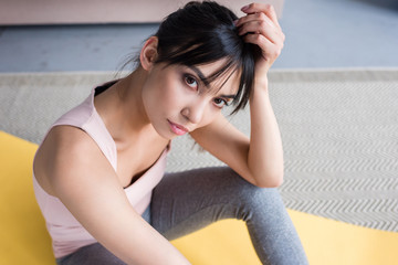 Fototapeta premium high angle view of beautiful young woman sitting on yoga mat and looking at camera at home