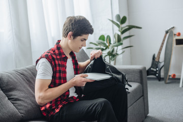 male teenager putting laptop into bag while sitting on sofa