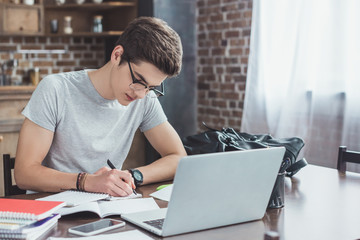 serious student writing homework at table with laptop and smartphone