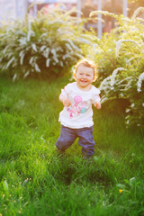  A funny baby girl takes the first steps and smiling at the bright green grass in the park in the summer.