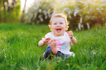  A funny baby girl is playing and smiling at the bright green grass in the park in the summer.
