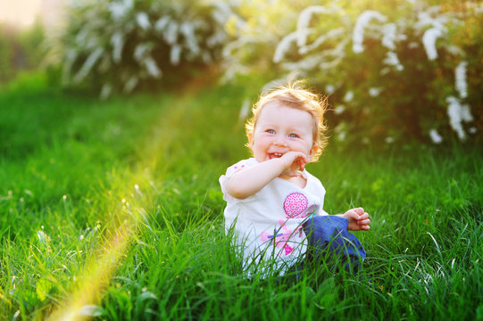 A funny baby girl is playing and smiling at the bright green grass in the park in the summer.