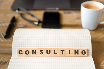 Closeup on notebook over wood table background, focus on wooden blocks with letters making CONSULTING word