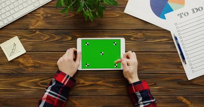 Close up of the female hands with the motley sleeves working on the white tablet device with a green screen horizontally on the wooden office table with a keyboard, papers, charts and a plant. Chroma