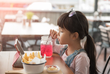 Beautiful laughing little girl sitting at table and eating French fries from your plate. See the iPad