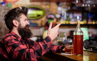 Hipster with beard ordered full bottle of alcohol. Relaxation concept. Man drinks whiskey or cognac. Man with happy face sits near bar counter. Guy spend leisure in bar, defocused background.