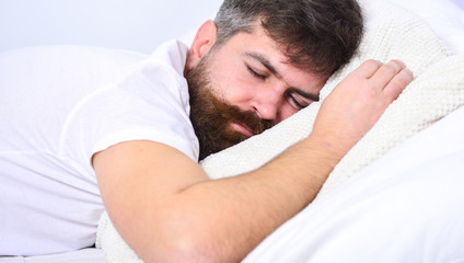 Man in shirt laying on bed, white wall on background. Macho with beard and mustache sleeping, relaxing, having nap, rest. Nap and siesta concept. Guy on calm face sleeping on white sheets and pillow.