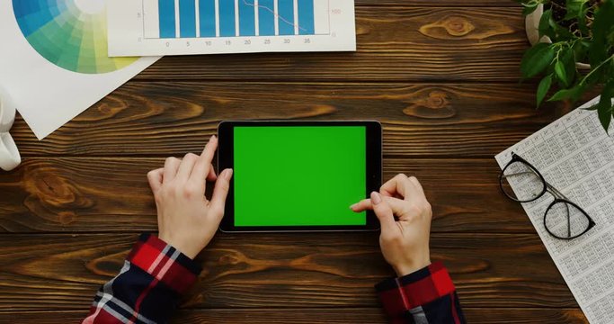 View from above on the female hands with the motley sleeves using the black tablet device with a green screen horizontally on the wooden office desk with papers, charts, glasses and a plant. Chroma