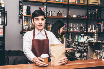 Handsome barista standing behind a counter and giving coffee cup to customer at cafe
