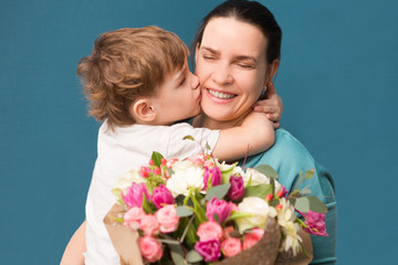 Fototapeta na wymiar Cute little child hugging and kissing her mother and offering flowers on a blue background. Mother's day, birthday