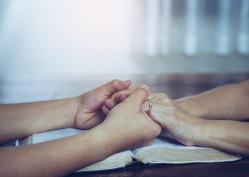 Two people are praying together over holy bible on wooden table