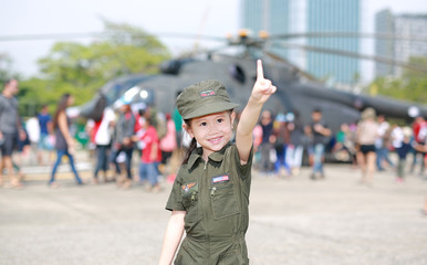 Smiling Asian child girl in pilot soldier suit costume with pointing up against blur helicopter background.
