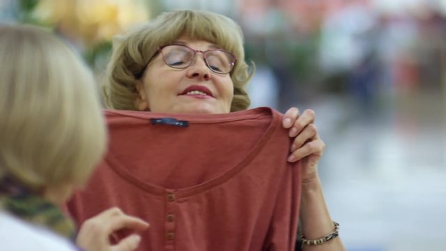 Medium shot of retired woman in glasses trying on long sleeve t-shirt when spending time with friends in cafe in shopping center