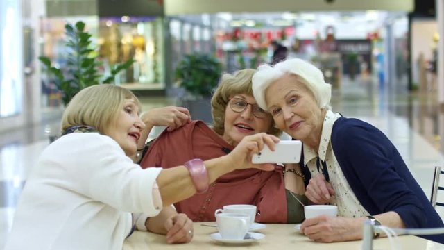 Medium shot of three cheerful retired ladies taking selfie on smartphone and laughing when sitting in cafe after shopping