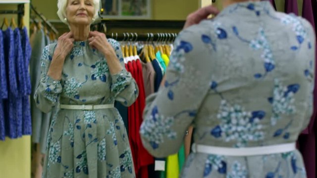 Tilt up shot of beautiful retired woman in pearl earrings trying on floral dress with white belt and posing in front of mirror