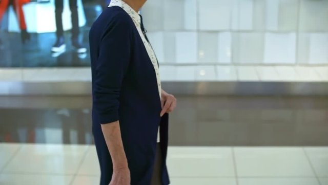 Side view of smiling old woman walking along shopping mall hallway and carrying paper bags with new clothes, tilt up shot