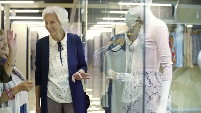 Tracking shot of group of three stylish retired women talking about clothes when standing by display window in shopping mall