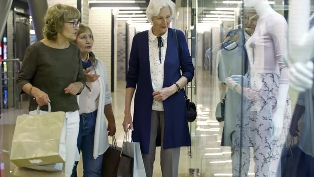 Group of three elderly women with paper bags stopping by display window of shopping center store and discussing outfits