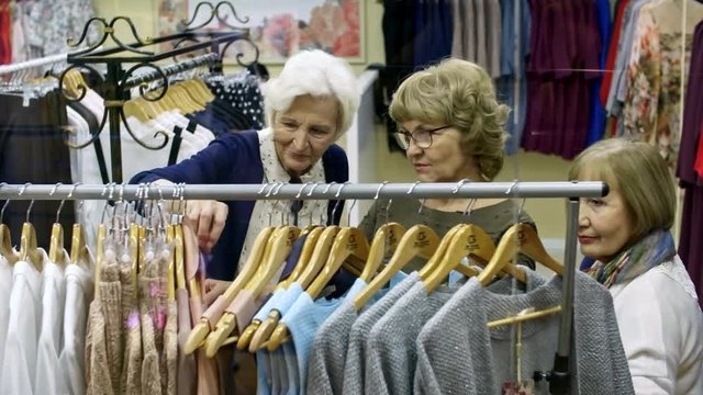 Group of three elderly female friends standing by clothing rack in shopping center, talking to each other and choosing outfits