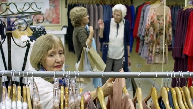 Medium shot of stylish retired woman standing by clothing rack in shopping mall and searching for outfits, two other women discussing blue dress in the background
