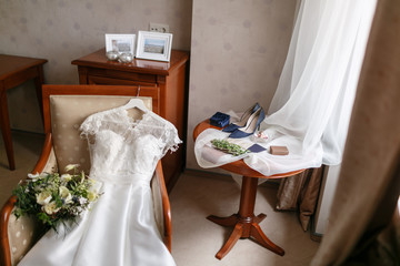 white wedding dress with lace lying on the chair. bouquet different flowers. Modern bridal bunch with poppy flower and thistle. bride accessories on the table