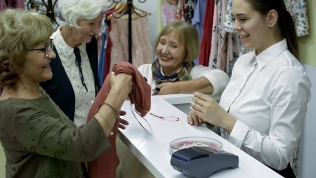 Medium shot of three retired women approaching cash desk in clothes store, one of them buying long sleeve t-shirt