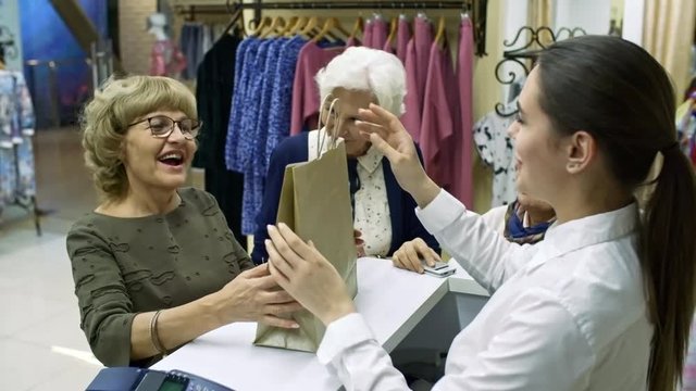 Medium shot of three elderly women standing at cash desk in clothes store, one of them taking paper bag from polite cashier