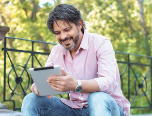 Cheerful adult man holding his digital tablet and surfing in the park.