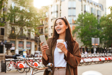 Woman outdoors with bicycle on the street chatting by mobile phone drinking coffee.