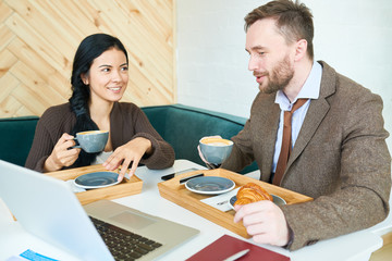 Portrait of two modern business people, man and woman, sitting at table smiling happily while drinking coffee with croissants and chatting  enjoying coffee break or lunch break during meeting in cafe