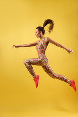 Fototapeta na wymiar Skinny girl with beautiful body, long legs, wearing colorful sportswear and sport shoes jumping high isolated on bright yellow background