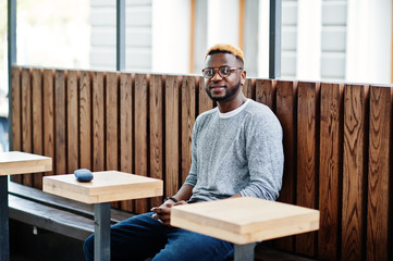 Stylish african american boy on gray sweater and glasses posed at street sitting on table wooden cafe. Fashionable black guy.