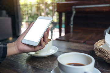 Mockup image of a woman's hand holding white mobile phone with blank desktop screen with coffee cup on wooden table in cafe