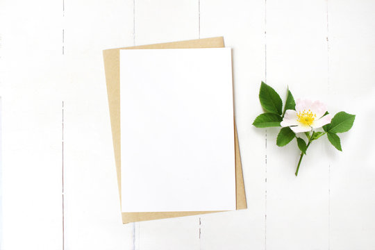 Feminine wedding stationery, floral desktop mock-up scene. Blank greeting card, craft envelope and blooming wild rose branch. Old white wooden table background. Flat lay, top view.