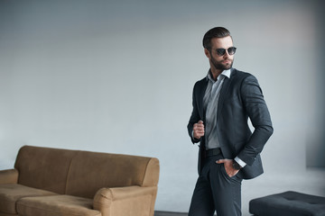 Young bearded stylish businessman leader indoors at office wearing sunglasses walking fixing sleeves with watch looking aside serious