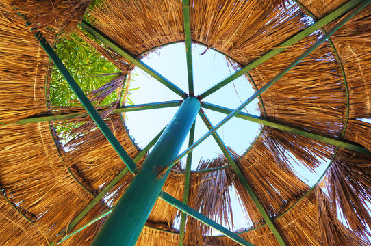 A beach umbrella made of peeled green metal and dry palm leaves from which one can see the sky and a green tree