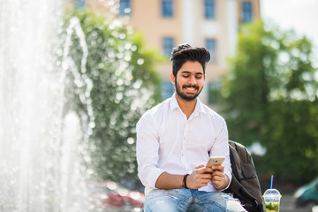 Young indian student man sitting on the street and look on the phone on fountains background