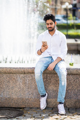 Young handsome indian man speak on phone sitting near fountain in the hot sunny summer day