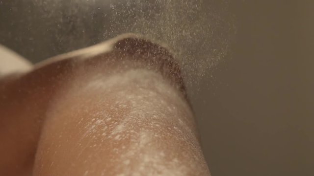 Naked skin of female leg bent at knee indoors. White cosmetic powder is flying from up to down and lying on foot, close-up