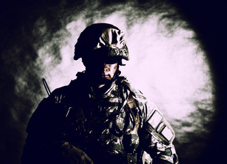 Low key studio portrait of private security service contractor, army infantry rifleman, US marine raider in helmet, sunglasses, camouflage uniform posing with weapon on black background with backlight