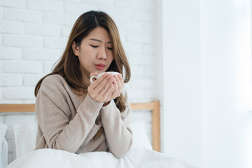 Beautiful Asian woman enjoying warm coffee on bed in her bedroom. Relaxation in bed. Beautiful Asian female wearing comfortable sweater holding a cup of coffee. lifestyle Asian woman at home concept.