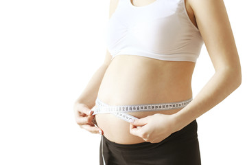 Close up of pregnant woman wearing supportive seamless maternity bra & black yoga pants measuring size changes of her bare tummy with tape measure flexible ruler. White background, copy space.