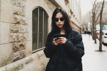 Portrait of cheerful asian hipster girl in trendy wear sending message via smartphone while standing on a street. Beautiful young woman in sunglasses using route application on mobile phone