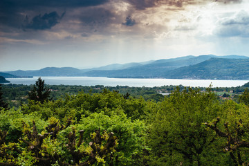 Fototapeta na wymiar panorama of Lake Maggiore green woods around and cloudy sky with rays of light, storm approaching