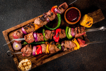 Fresh, home-cooked on the grill fire meat beef shish kebab with vegetables and spices, with barbecue sauce and ketchup, on a dark background on a wooden cutting board above copy space - obrazy, fototapety, plakaty