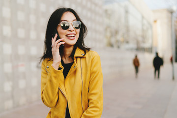 Smiling asian woman with long dark hair in sunglasses talking by a smartphone while walking the city street. Young entrepreneur making business calls by a mobile phone while going to coworking space. - 205887054