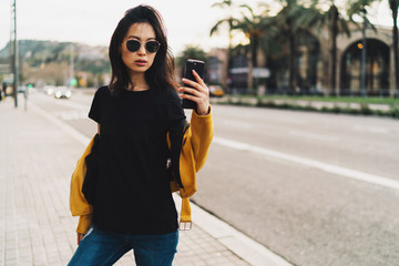 Fototapeta premium Stylish asian woman with long dark hair in sunglasses wearing blank black t-shirt and yellow leather jacket taking selfie by a mobile phone while standing on a blurred urban background.
