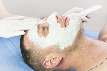 A man does the procedure cleaning his face with a clay mask in the beauty salon 