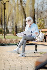 Interesting news. Unsmiling aged woman reading a newspaper while sitting on the bench